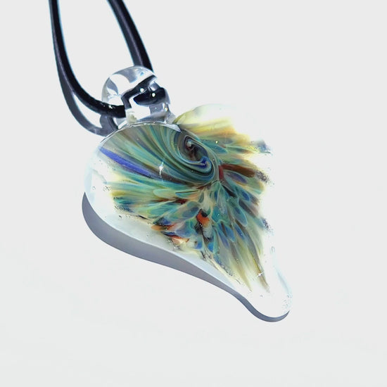 Heart-shaped handmade glass pendant with psychedelic swirl design in blue, green, and amber, suspended on a black cord against white background.