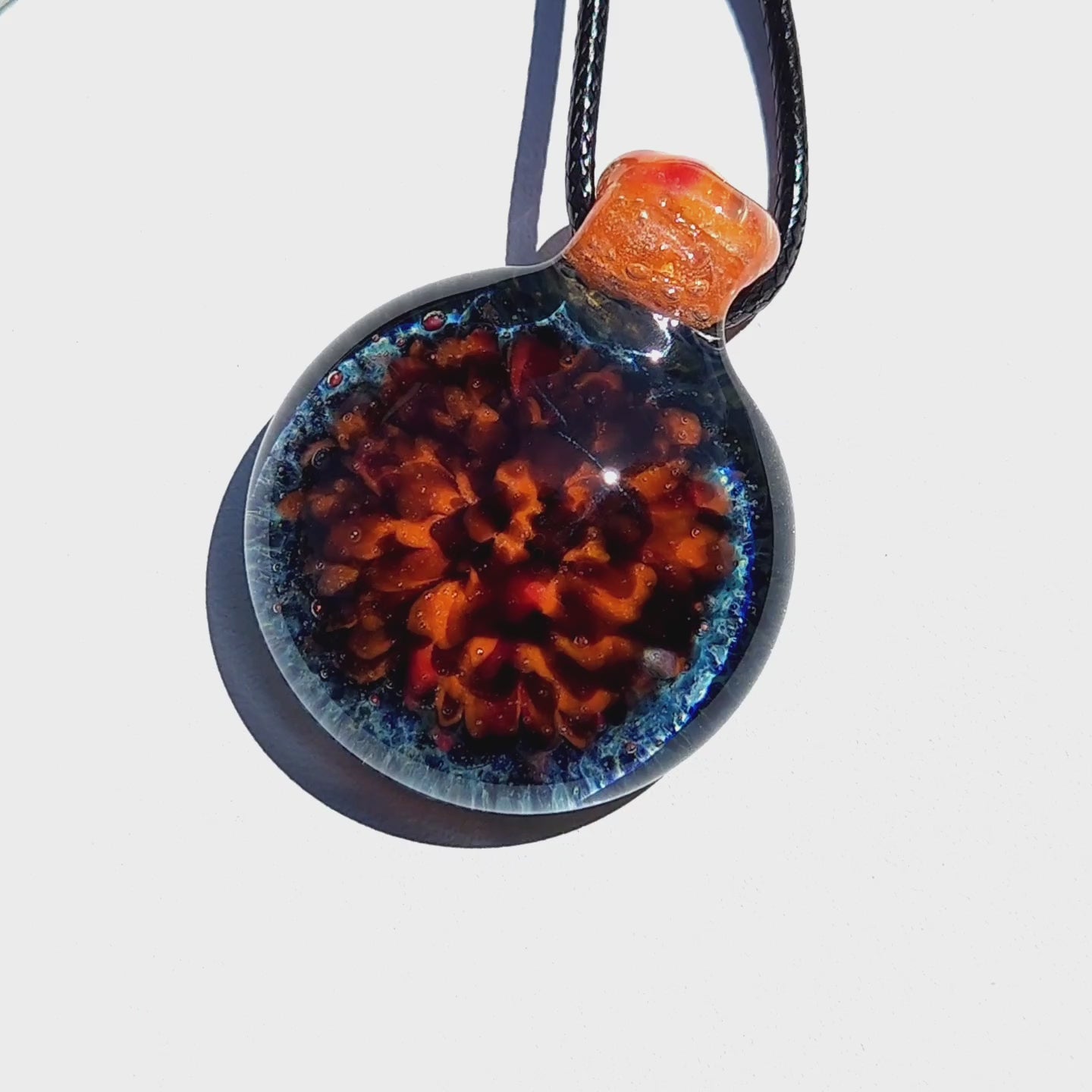 Round handmade glass pendant with vibrant orange core and blue rim, suspended on black cord. One-of-a-kind borosilicate jewelry necklace.