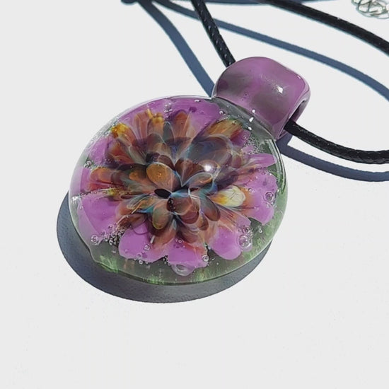 Angled view of heady glass pendant with intricate flower encased in clear glass. Handmade borosilicate necklace in purple and green hues.